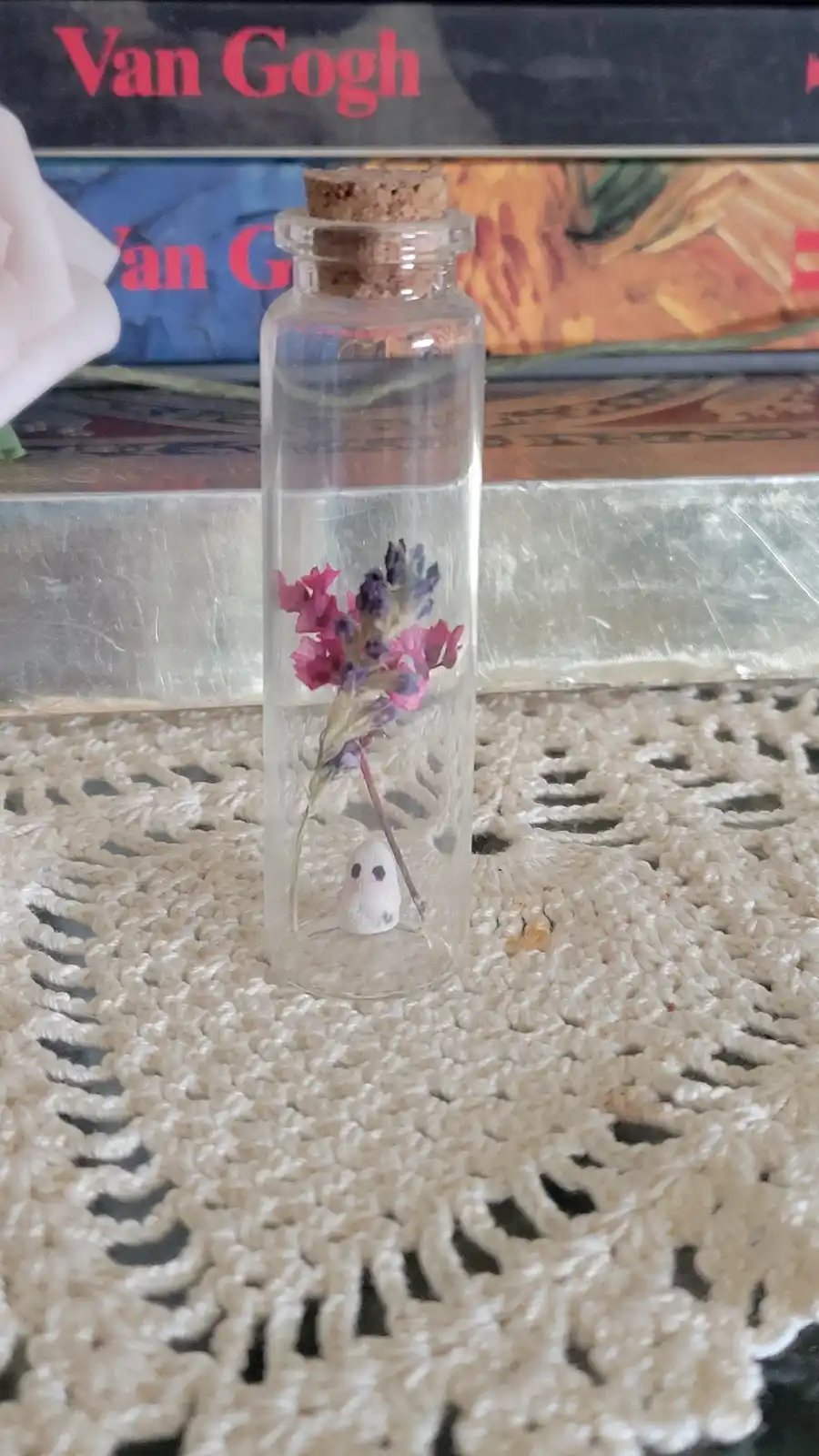 A small glass bottle with a cork stopper sits on a white crocheted doily, containing a tiny white ghost figurine and a dried…