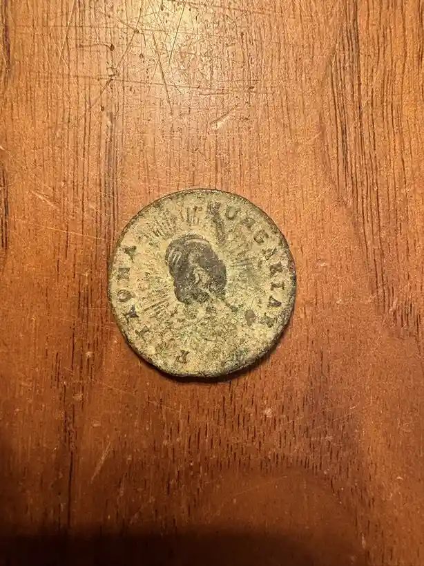 A close-up photo shows a weathered, circular ancient coin with a profile bust and Latin text, resting on a scratched wooden…