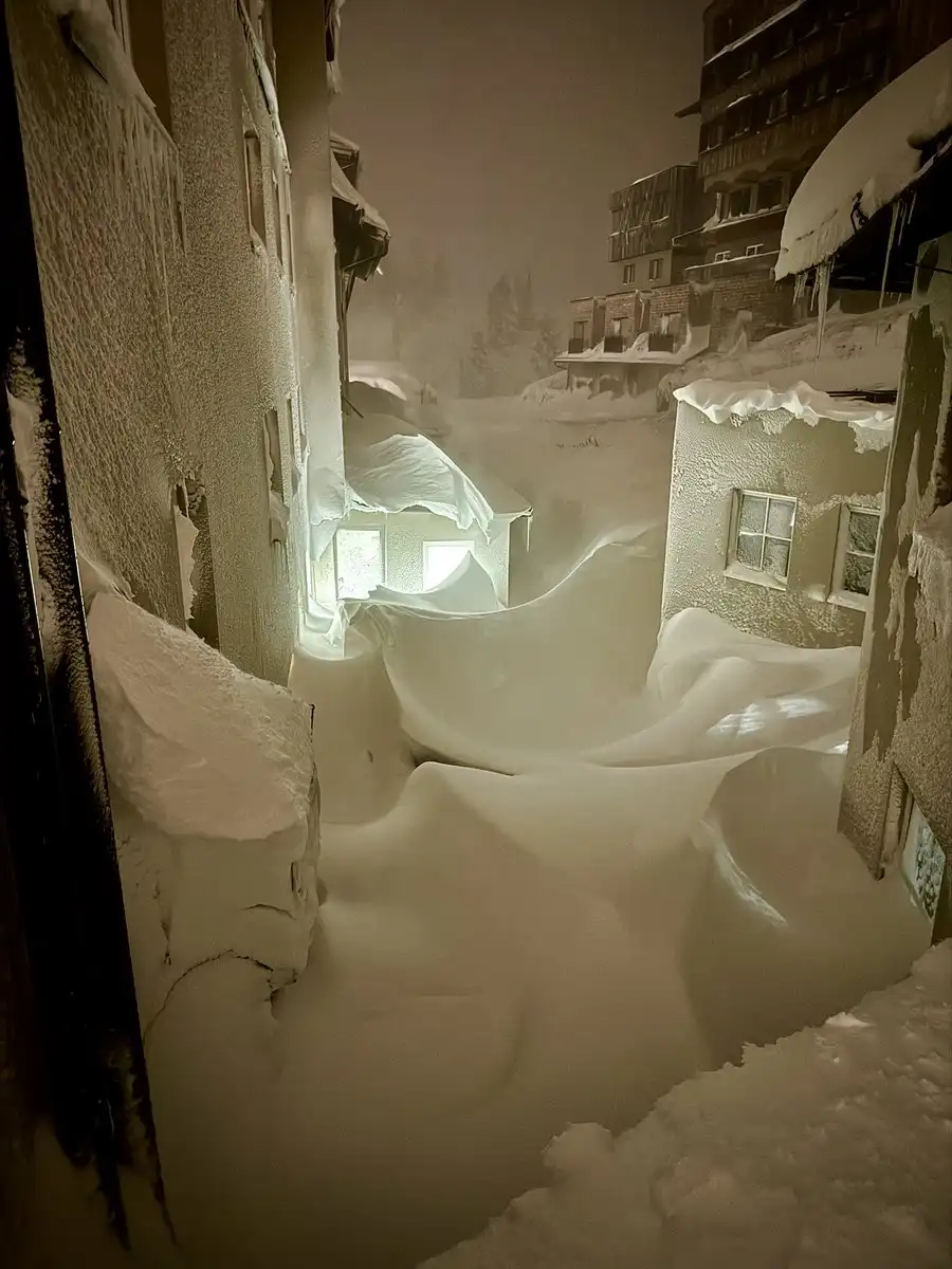 A nighttime photo shows a narrow alleyway between buildings completely buried under deep, sculpted snowdrifts, with only a…