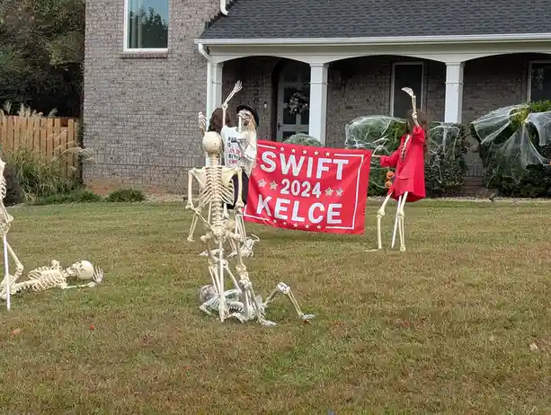 Several Halloween skeleton decorations are arranged in a yard in front of a brick house, holding a red banner that reads…