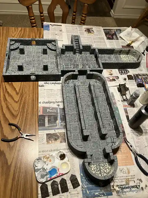 A tabletop view of a partially painted miniature dungeon set, laid out on newspaper with paint supplies, pliers, and small…