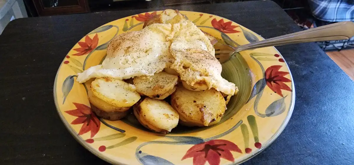A plate of fried eggs and roasted potato slices sits on a yellow floral-patterned plate with a fork resting on the side. The…