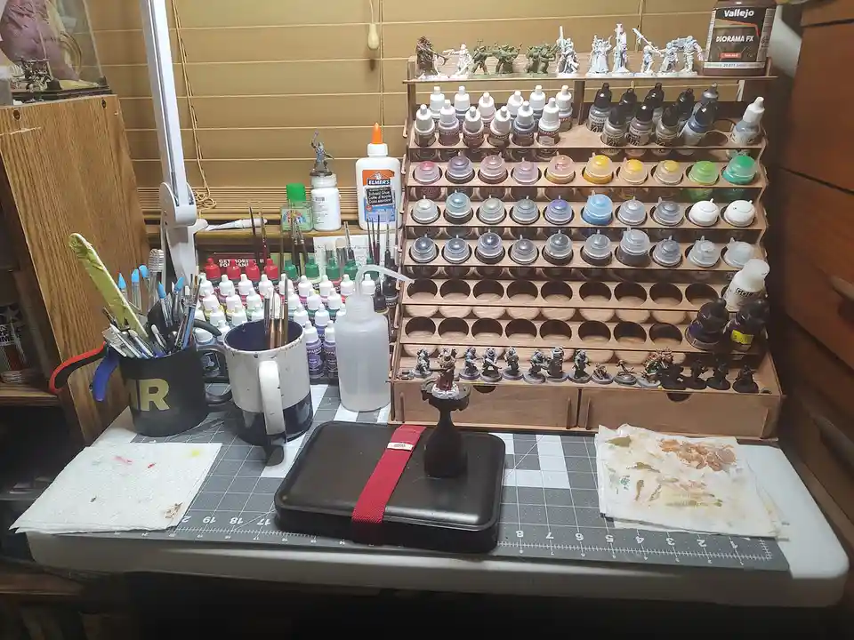 A cluttered hobbyist's desk is filled with miniature painting supplies, including numerous paint bottles organized in a…