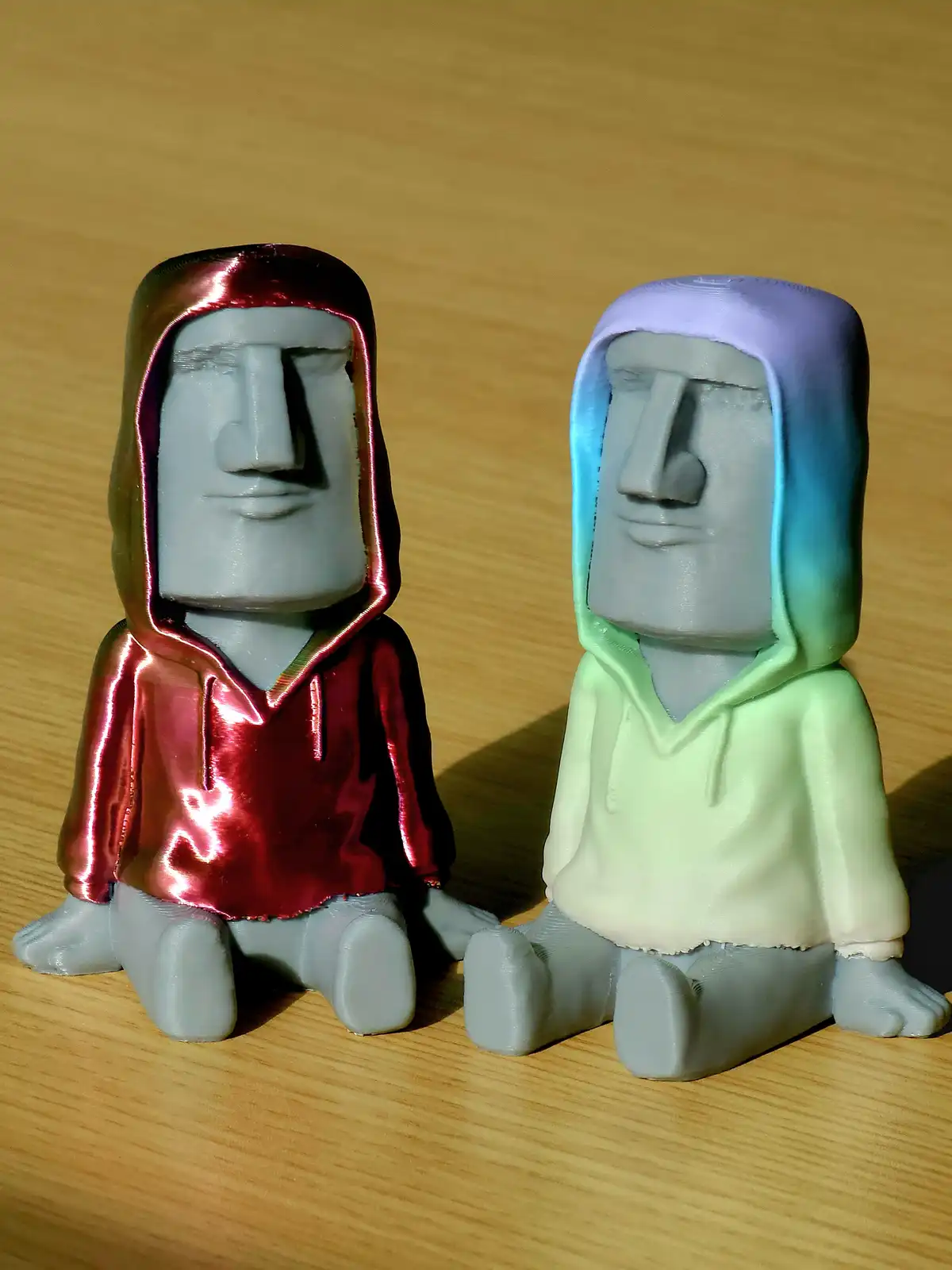 Two 3D-printed Moai statues sit side-by-side on a wooden surface, each wearing a hoodie. The left Moai wears a shiny red…