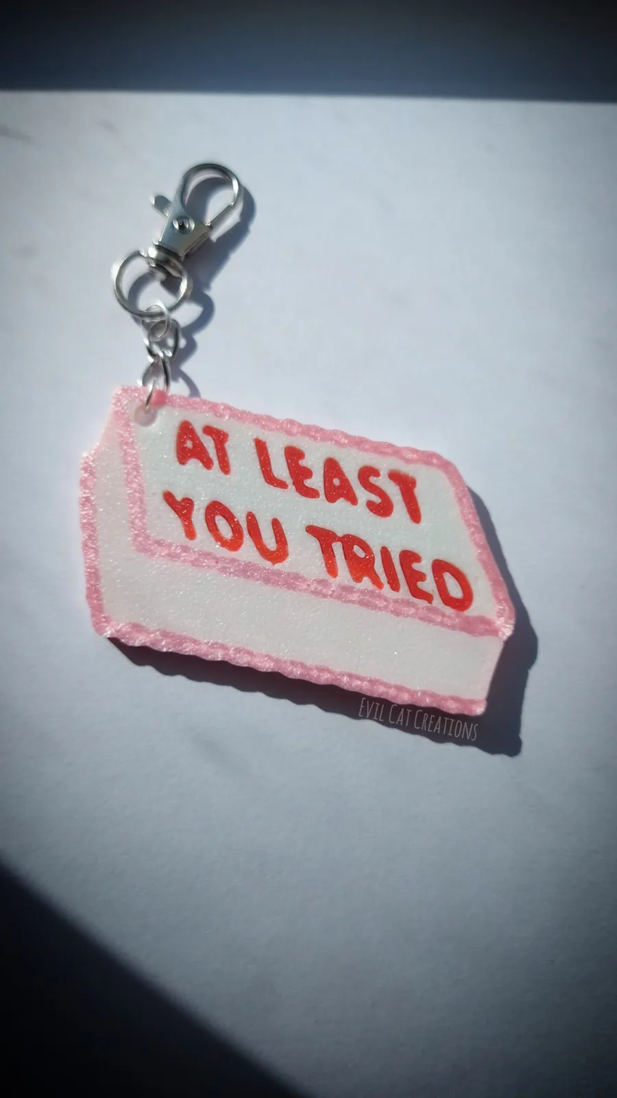 A handmade keychain with a white, glittery tag edged in pink, attached to a silver lobster clasp. The tag has red, glittery…