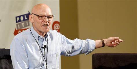 A bald man with glasses, wearing a light blue button-down shirt and a lanyard, is captured mid-speech, pointing emphatically…