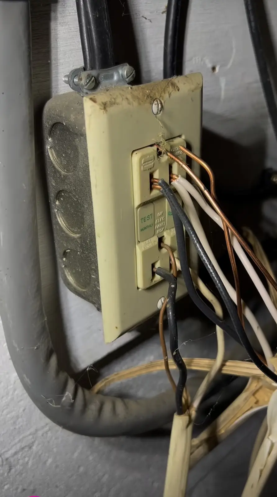 A close-up of a dirty, beige GFCI outlet with exposed copper and black/white wires crudely connected to its terminals…