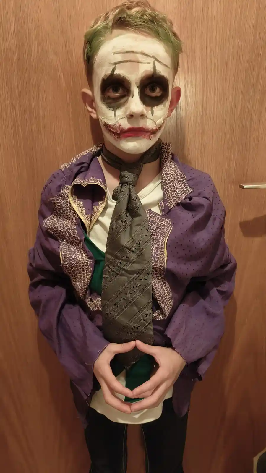 A young boy dressed as the Joker from Batman, with white face paint, black eye makeup, green hair, and a purple jacket with…
