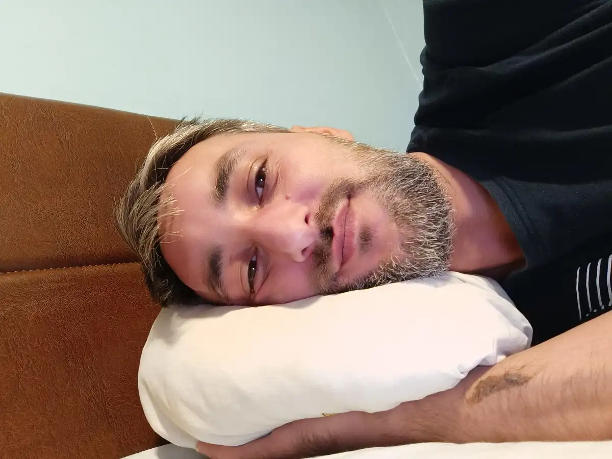 A man with a graying beard and mustache lies on his back on a brown leather couch, resting his head on a white pillow. He…