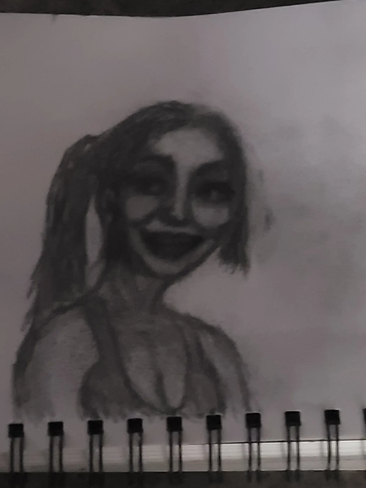 A blurry black-and-white sketch of a person with pigtails and a wide, unsettling grin, drawn in a sketchbook with visible…
