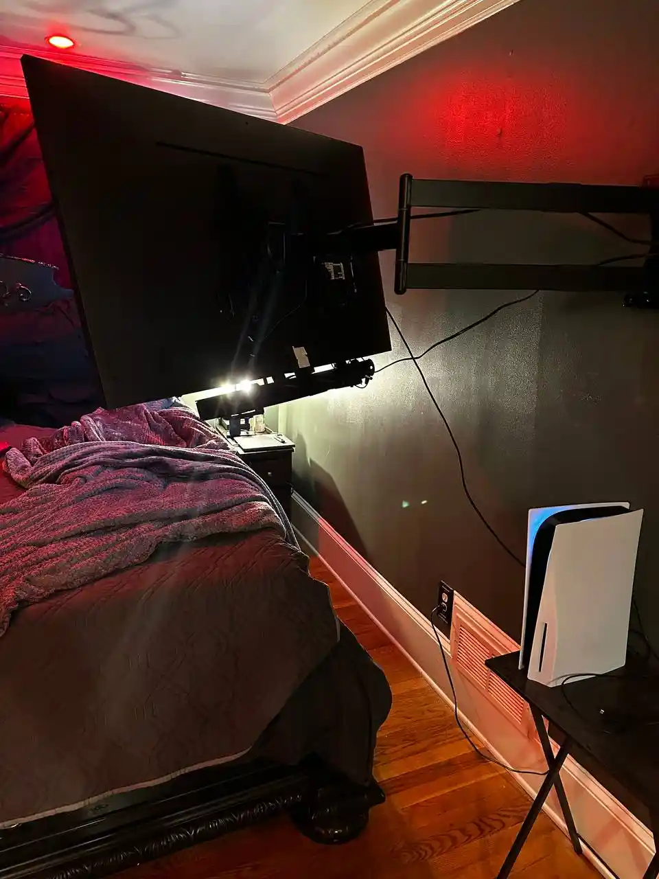 A bedroom scene with a large TV mounted on the wall, angled downward toward a bed with a purple blanket. A white PlayStation…