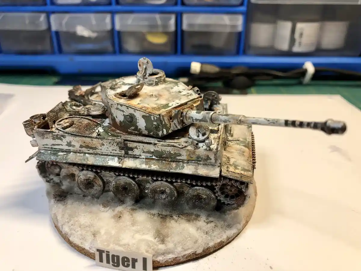 A highly detailed, weathered scale model of a Tiger I tank sits on a snowy base, showing chipped paint, rust, and battle…