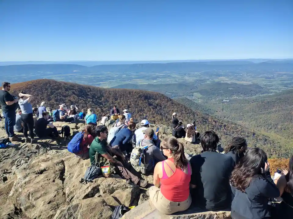 A large group of hikers sits on a rocky mountain summit overlooking a vast valley with rolling hills and autumn-colored…