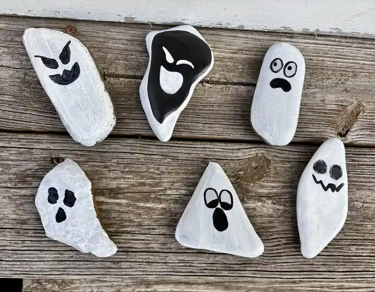 Six hand-painted rocks, each shaped and decorated to resemble a different cartoonish ghost with unique facial expressions…