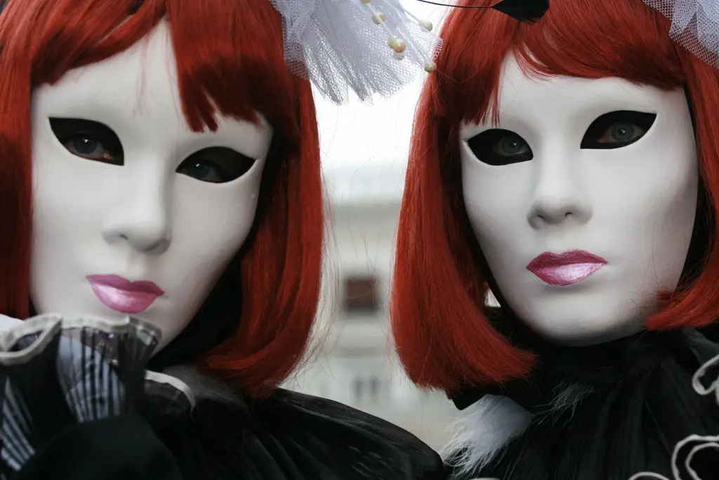 Two people in identical Venetian-style white masks with black eye cutouts and red wigs stand side by side, both wearing…