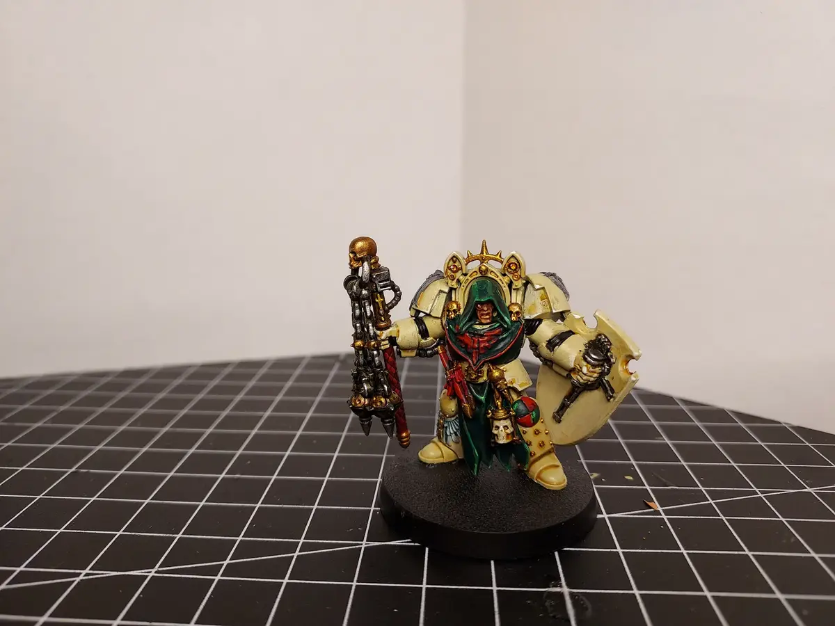 A painted miniature of a heavily armored, hooded warrior with a skull-topped weapon and shield stands on a black grid mat…