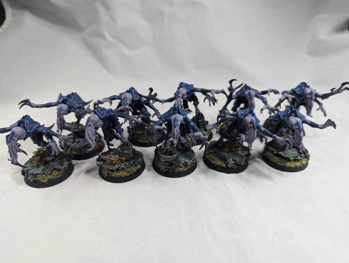A row of ten identical, intricately painted miniature figurines depicting grotesque, multi-limbed alien creatures with blue…