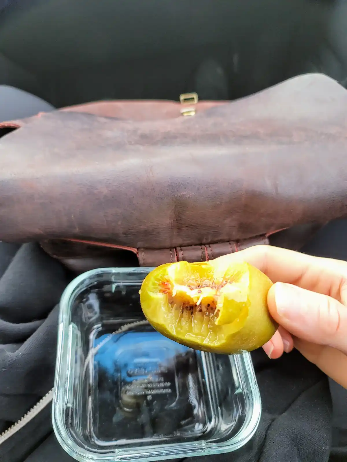 A hand holds a half-eaten green fig over an empty glass container, with a brown leather bag in the background. The overlaid…