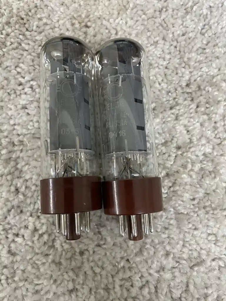Two vintage EL34 vacuum tubes with brown bases sit side-by-side on a white carpet. The tube on the left is labeled 'EL34…