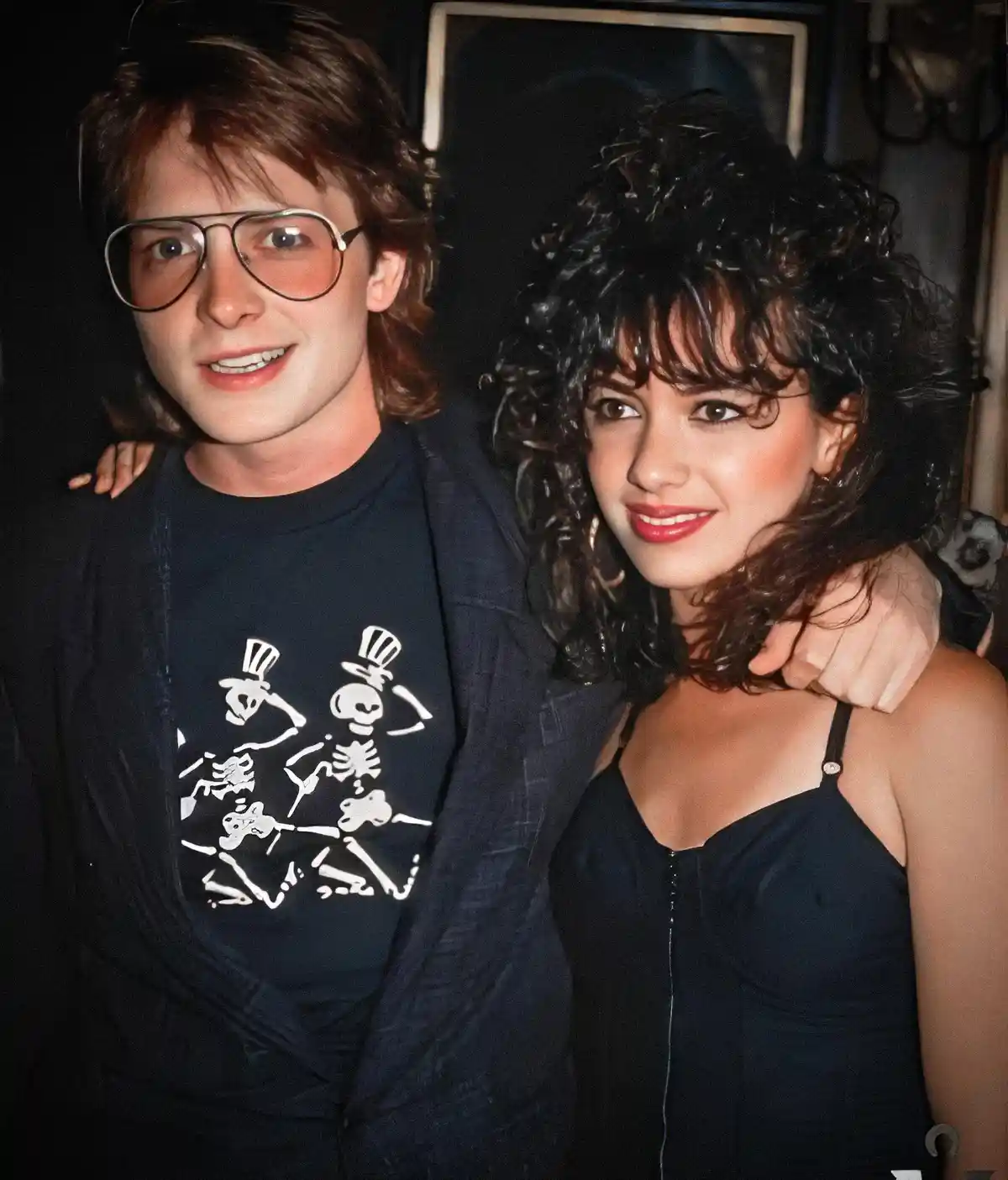 A man with 80s-style hair and large aviator glasses wears a black t-shirt with a white graphic of dancing skeletons in top…
