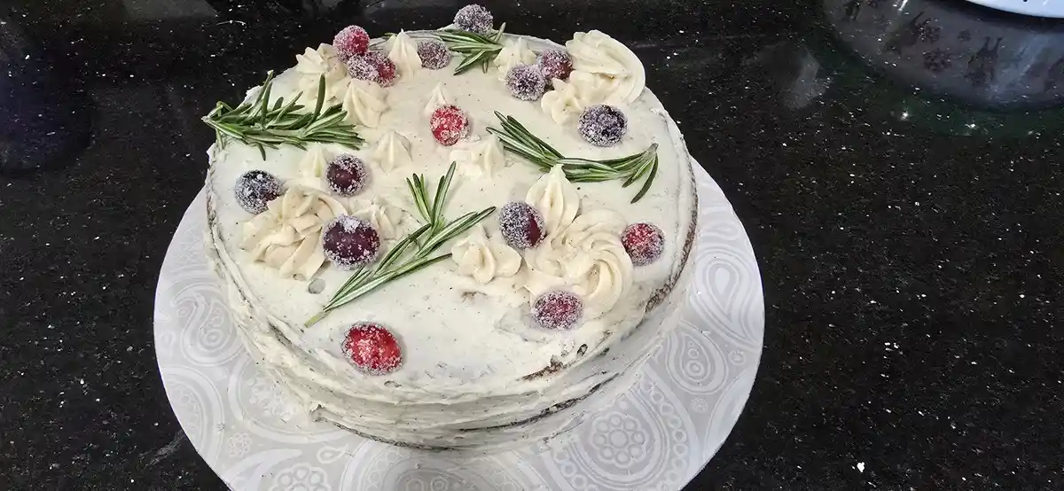 A round cake with light-colored frosting, decorated with sugared cranberries and sprigs of rosemary, sits on a patterned…