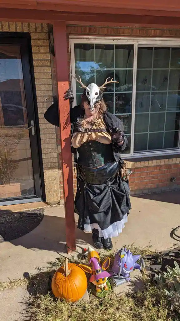 A person in a Halloween costume stands outside a brick house, wearing a deer skull mask with antlers, a corset, and a…