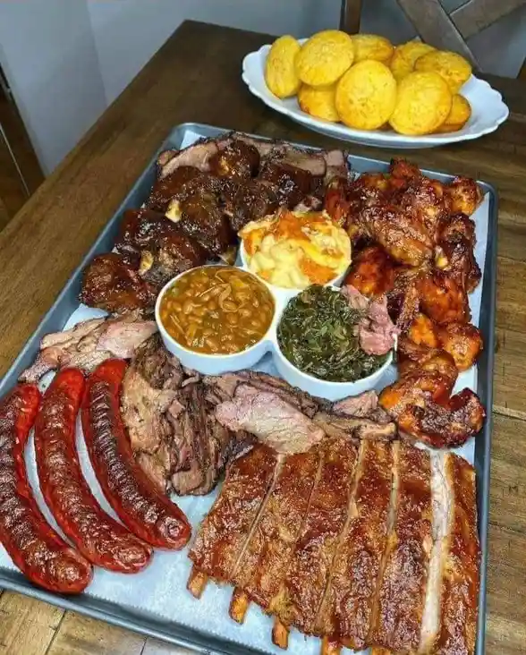 A large metal tray is filled with an abundant spread of barbecue food, including ribs, pulled pork, smoked sausages, chicken…