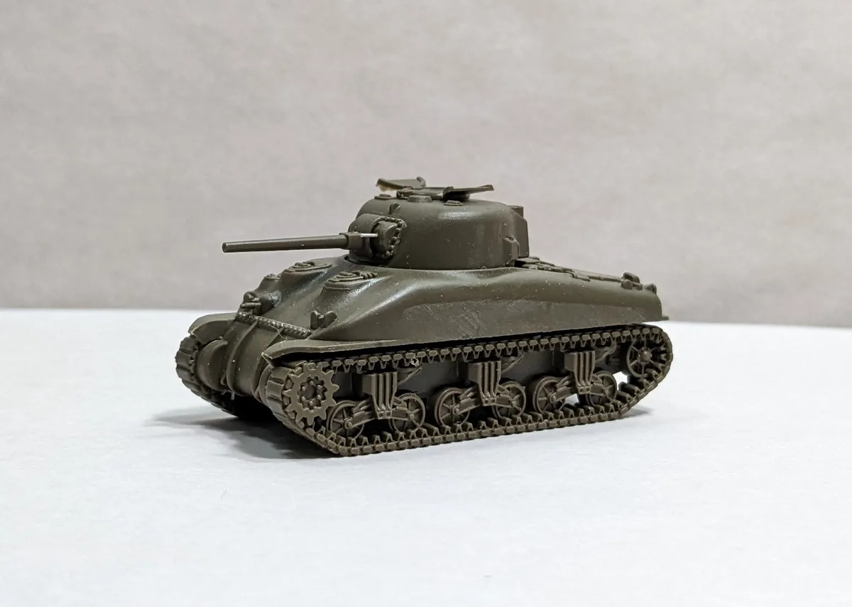 A small, olive-drab model of a military tank sits on a plain white surface against a neutral gray background. The tank has a…