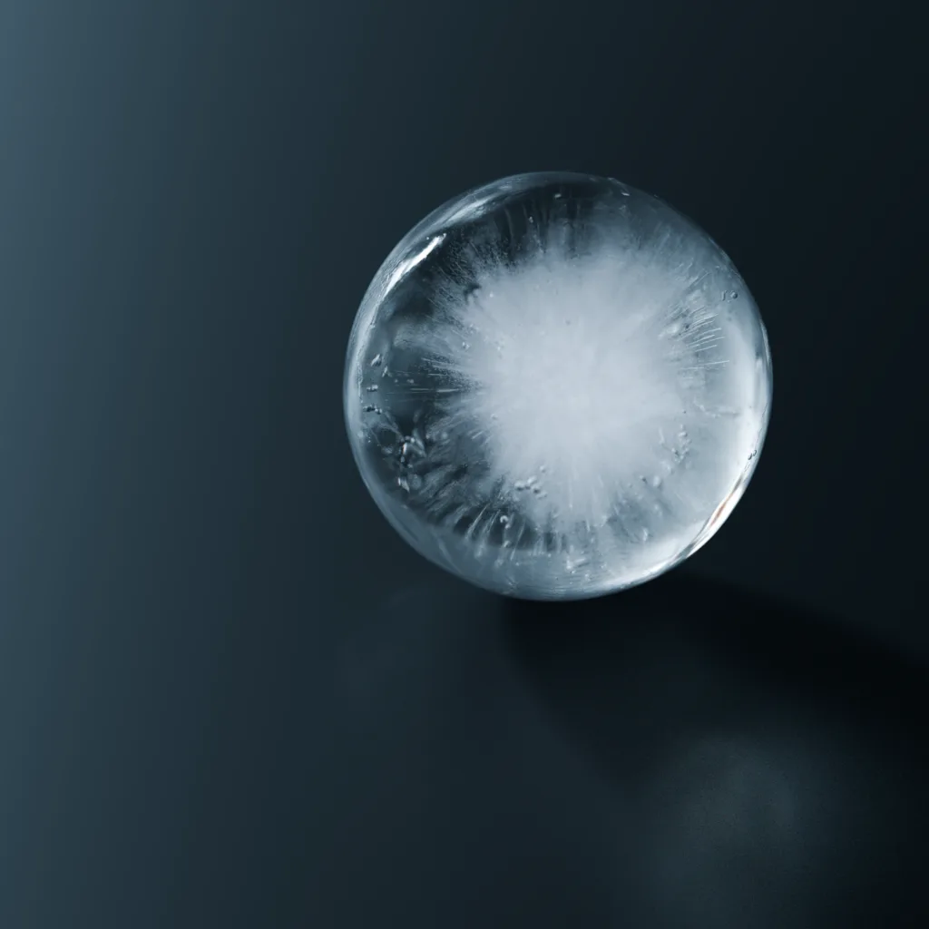 A close-up photograph of a clear, spherical ice ball resting on a dark, reflective surface. The ice has a crystalline…