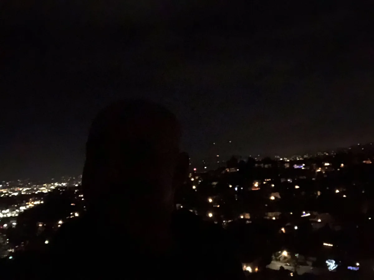 A dark silhouette of a person is centered in the foreground, facing forward, against a nighttime cityscape backdrop filled…