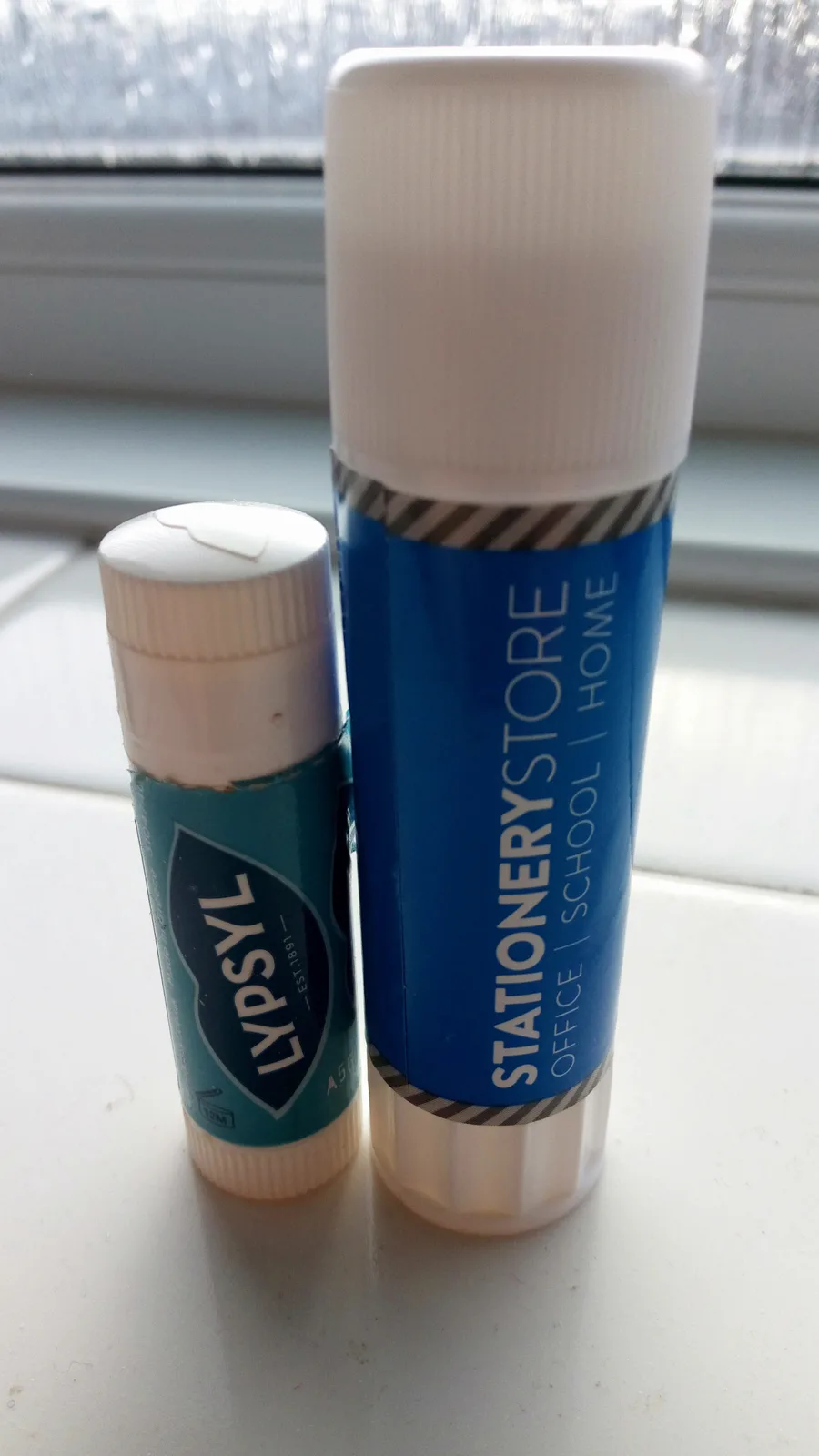 Two glue sticks sit side by side on a windowsill, one small and one large. The small one has a teal label with 'LYPSYL'…