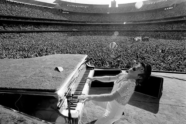 A black-and-white photo shows a man in a sparkly outfit and sunglasses, leaning back while playing a grand piano on a stage…