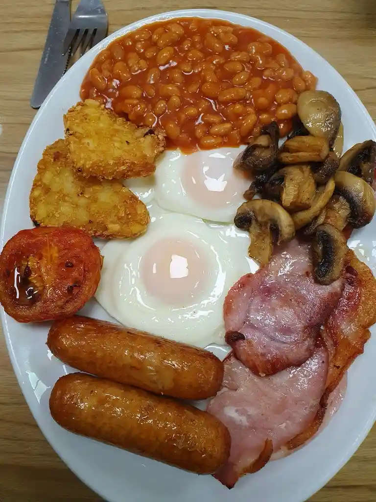 A plate of full english breakfast with baked beans, fried eggs, hash browns, mushrooms, bacon, ham, sausages, and a grilled…