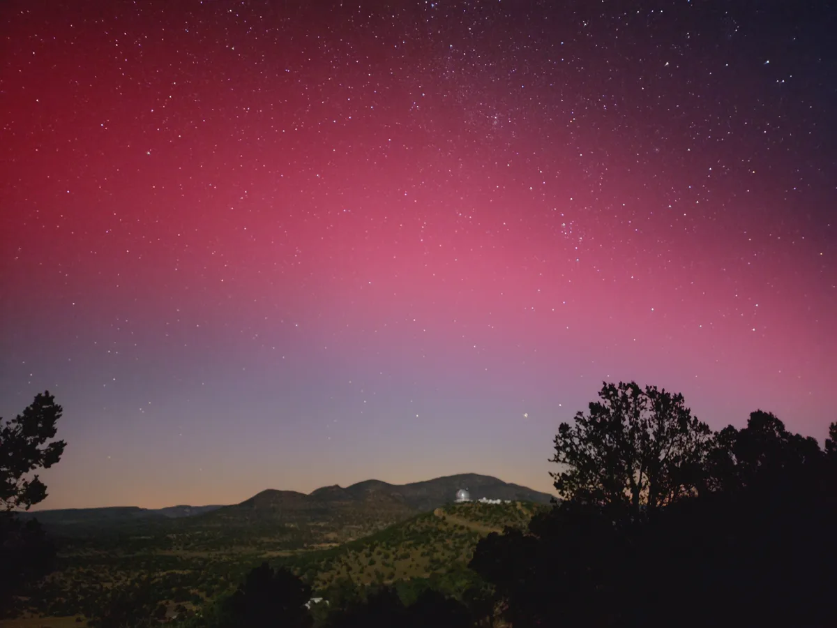 A night sky photograph shows a vibrant pink and purple gradient filled with countless stars, above silhouetted hills and…