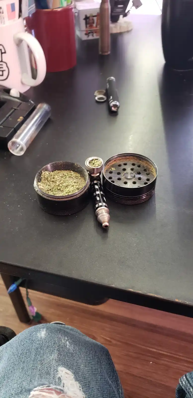 A black two-part herb grinder sits open on a dark table, its bottom half filled with ground green plant material and its top…