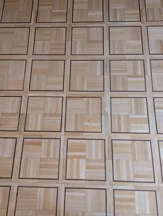 A close-up photo of a light-colored wooden parquet floor with dark grout lines forming a grid pattern. One square in the…