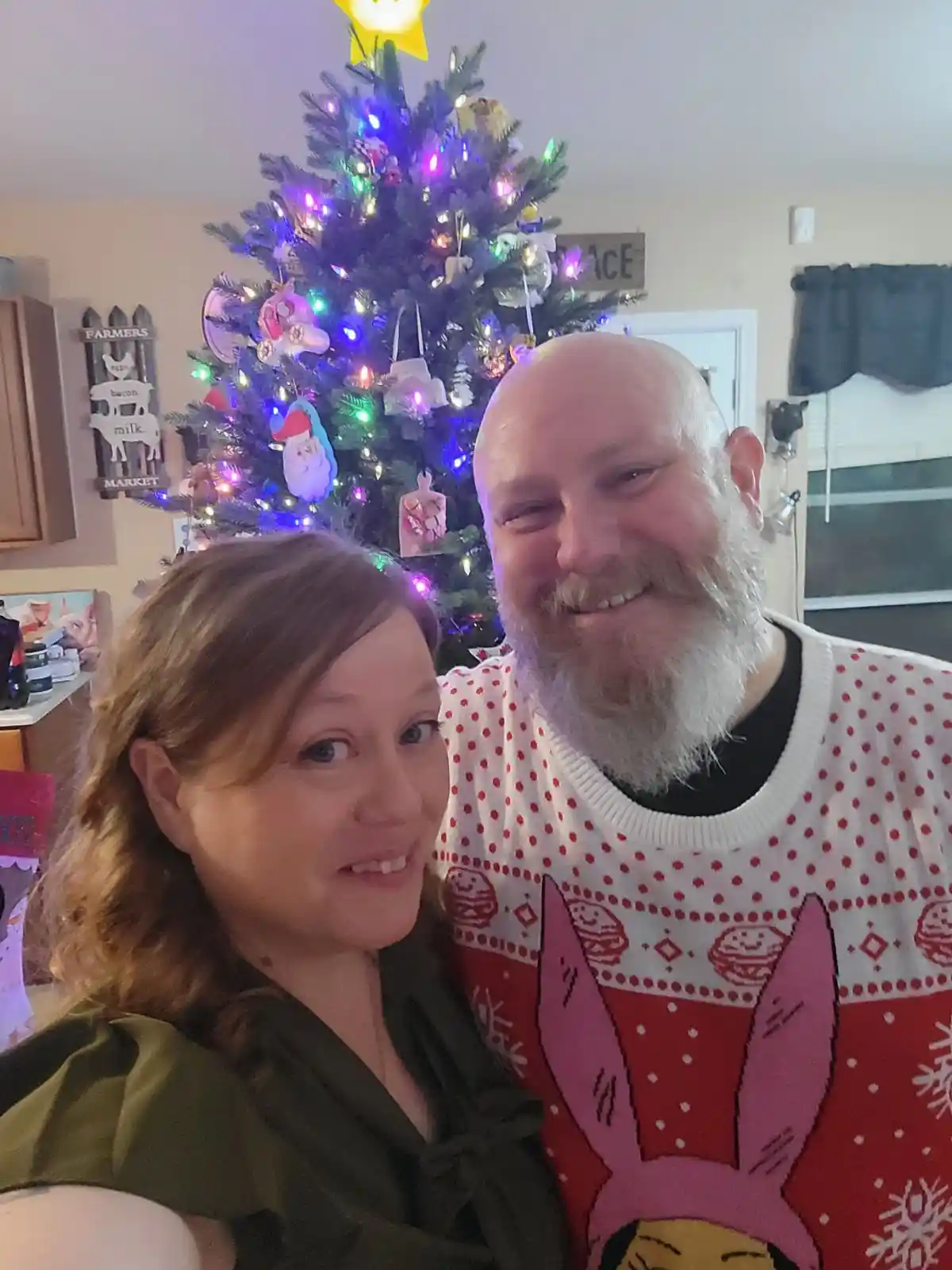 A man with a white beard and a woman are posing for a selfie in front of a decorated Christmas tree. The man is wearing a…