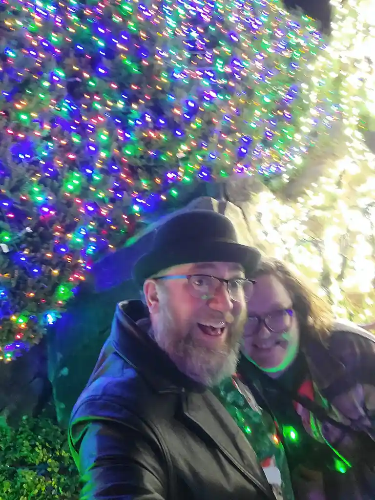 A blurry selfie of a man and woman smiling under a canopy of colorful, blinking holiday lights. The man wears a black hat…