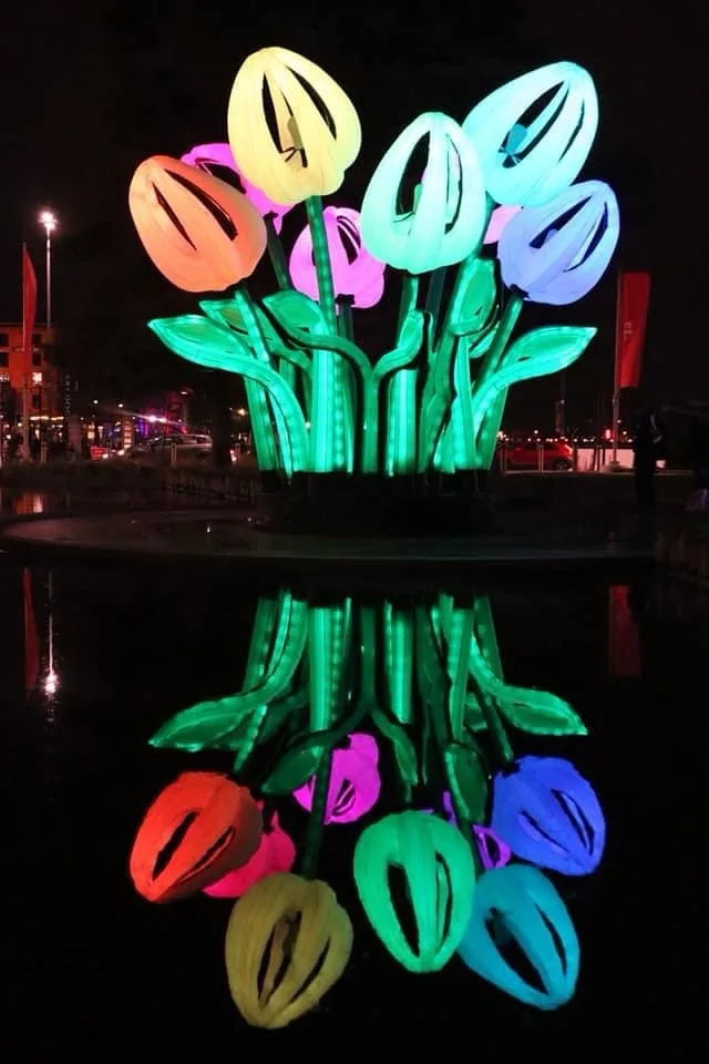 A large, illuminated sculpture of colorful tulips stands in a dark pond at night, with its vibrant reflection clearly…