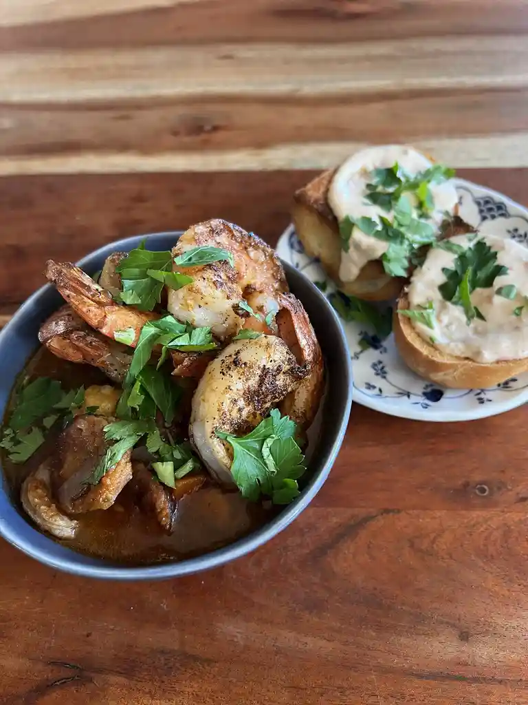A close-up photo shows a dark bowl filled with saucy shrimp and what appears to be grits or rice, garnished with chopped…
