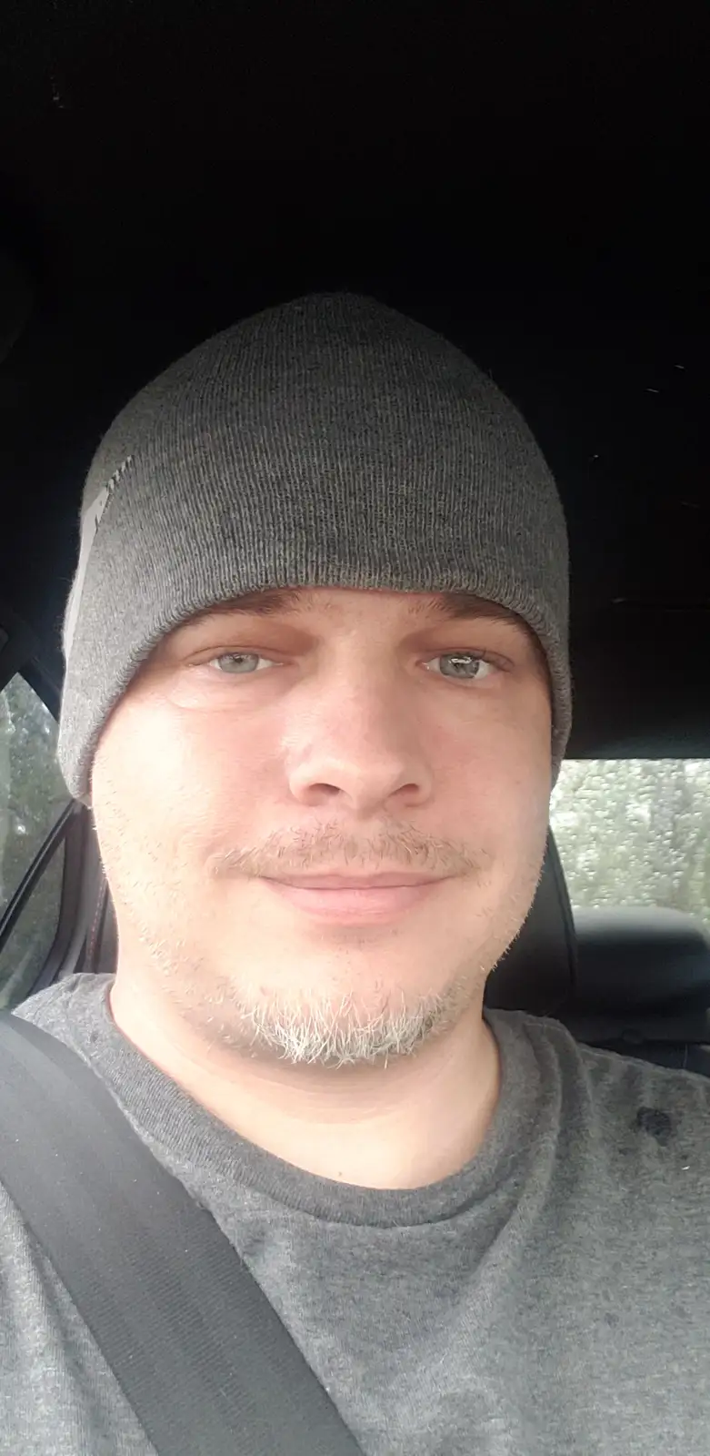 A man wearing a gray beanie and gray t-shirt sits in a car, looking at the camera with a slight smile. Raindrops are visible…