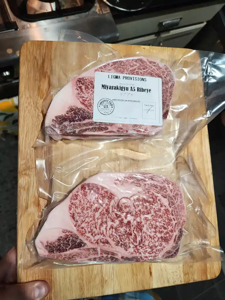 Two vacuum-sealed, high-grade Miyazakigyu A5 ribeye steaks sit on a wooden cutting board, their labels humorously altered to…