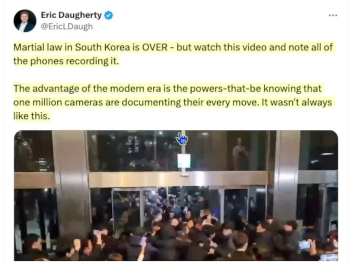 A nighttime photo shows a crowd of people gathered outside a building, many holding up phones to record the scene. A blue…