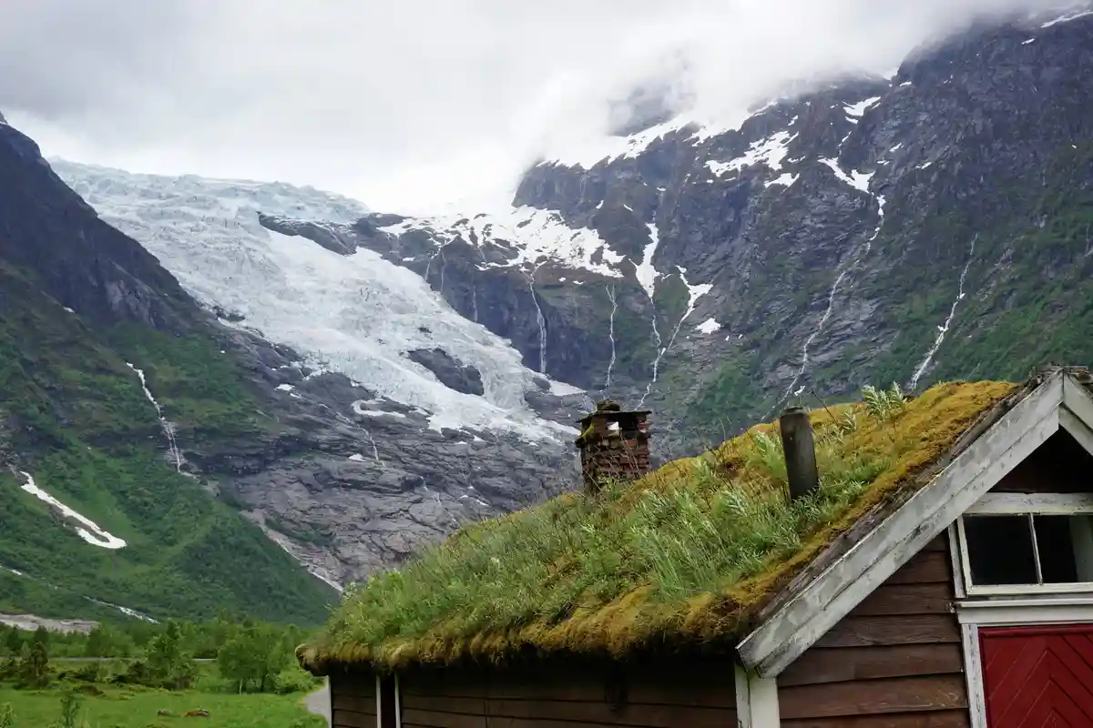 A rustic wooden house with a moss-covered roof and brick chimney sits in the foreground, with a massive glacier and…