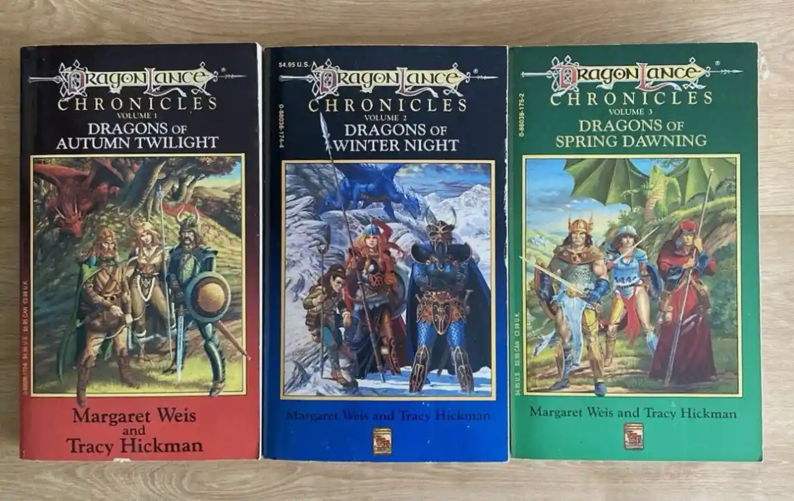 Three Dragonlance Chronicles paperback books, volumes 1-3, are lined up on a wooden surface. Each cover features fantasy…