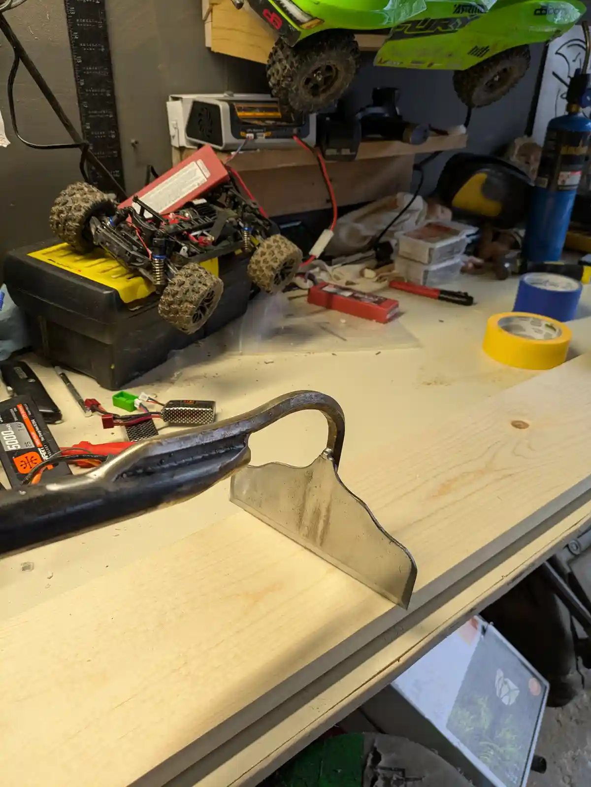 A cluttered workshop desk holds a custom-made metal tool resting on a wooden plank in the foreground. Behind it, a…