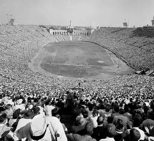 a black-and-white photo of a massive, packed baseball stadium with thousands of spectators filling the stands around a…