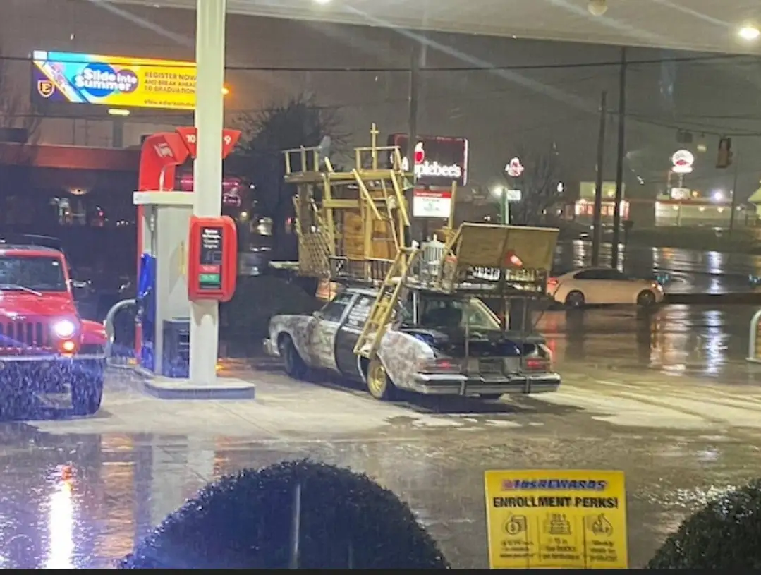 A heavily modified, camouflaged car with multiple ladders and scaffolding strapped to its roof is parked at a gas station at…