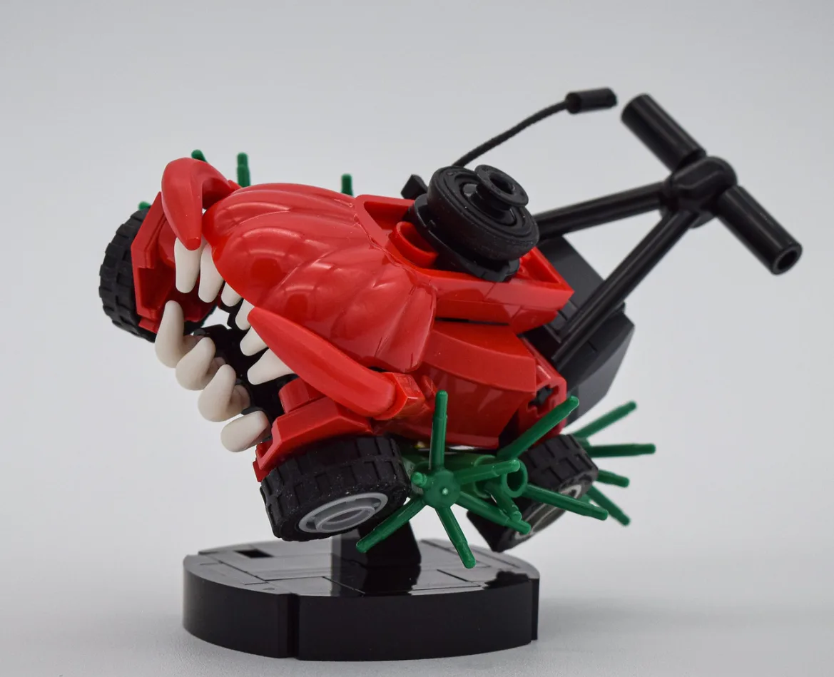 A red plastic toy lawn mower with a large, open mouth full of white teeth, green spiky blades, and black wheels, mounted on…