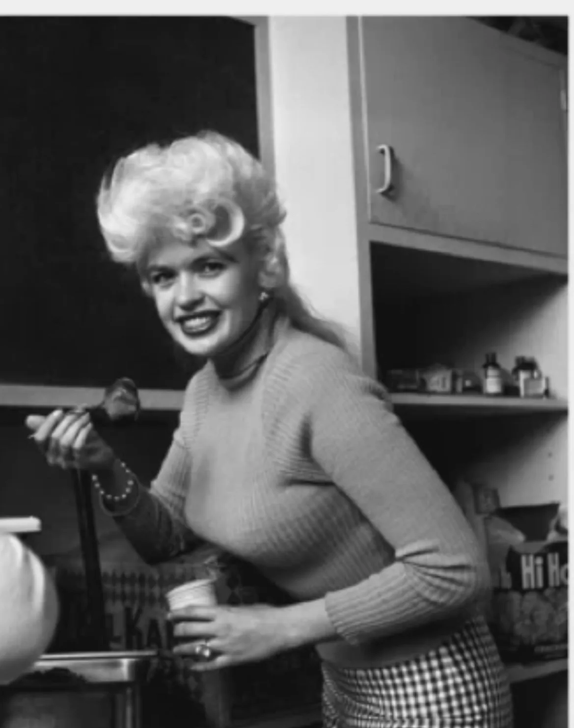 A black-and-white photo of a smiling blonde woman in a 1950s-style kitchen, wearing a ribbed sweater and checkered skirt…